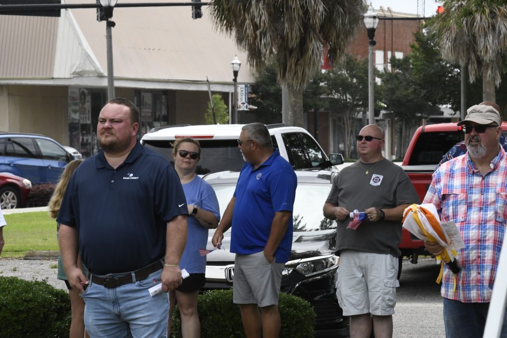 A group of people standing outdoors on a street near parked cars, with trees and buildings visible in the background on a sunny day.