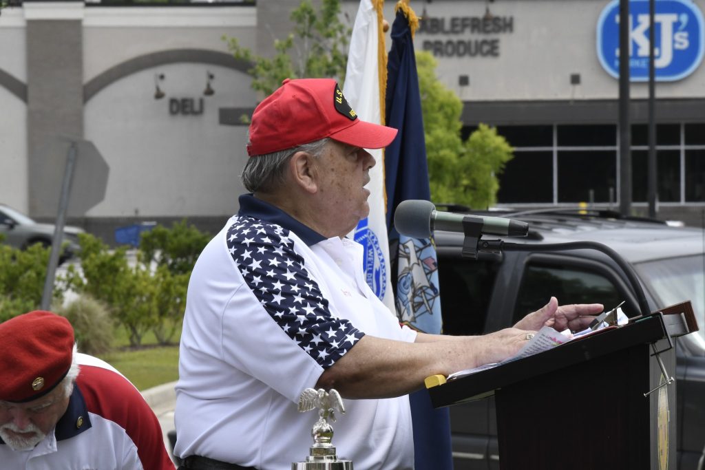 elderly man in a patriotic shirt and red cap speaks at an outdoor podium, with flags and a building visible in the background.