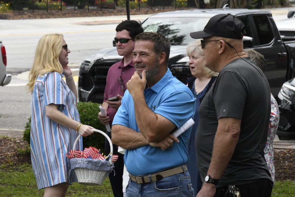 A group of people gathered outdoors, conversing and smiling; one woman holds a basket of items, and others are dressed casually.