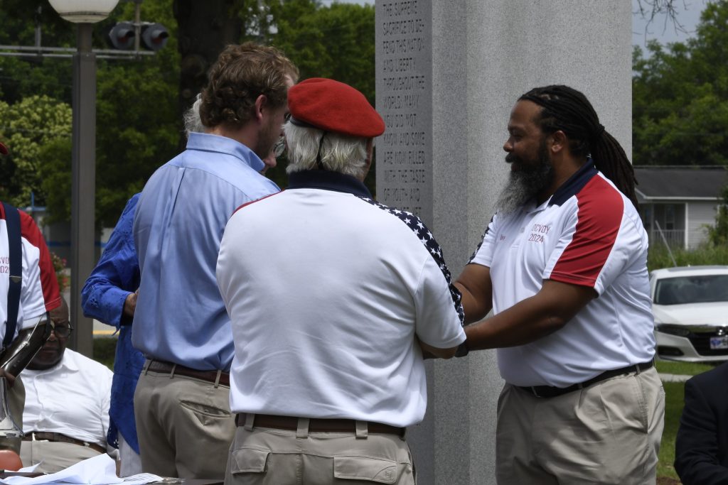 Four men stand together outdoors, engaged in conversation; one man wears a red beret, another wears a white shirt with red and blue sleeves.