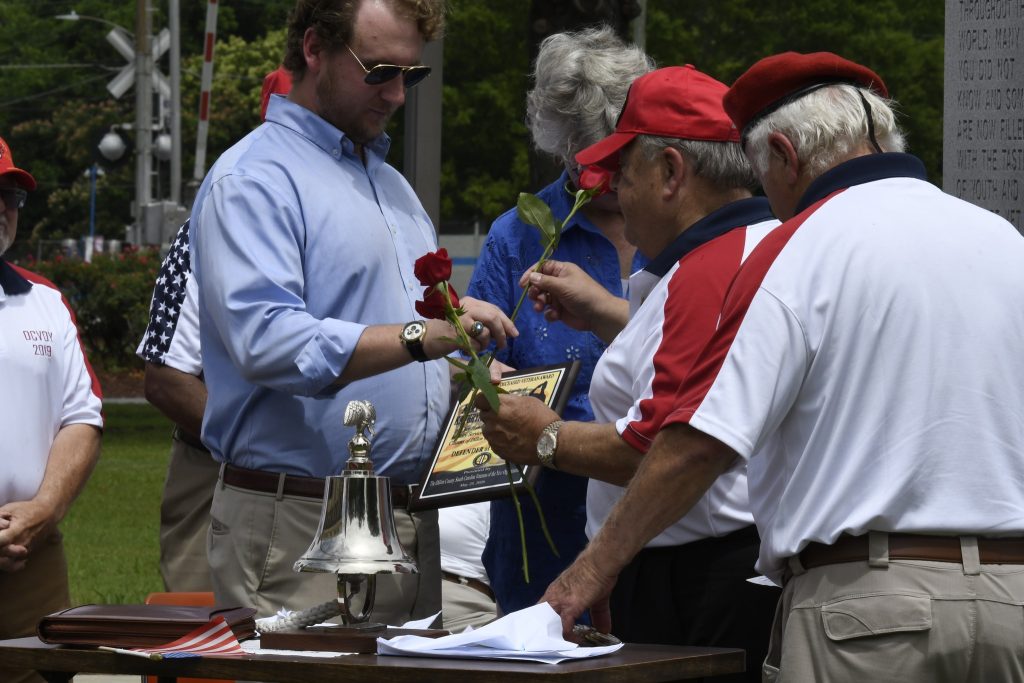 A group of people gathered outdoors, some holding roses and documents, engaging in conversation around a table with a bell and papers.