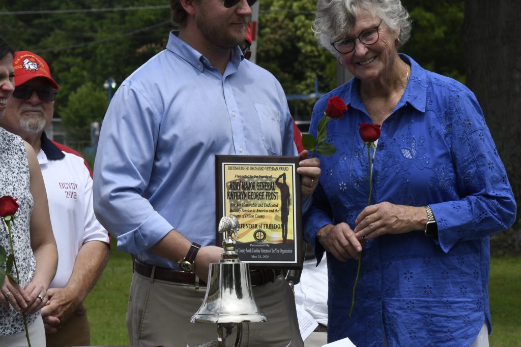 A man holding a plaque and a silver bell stands next to a woman in blue holding a red rose, both at an outdoor event.