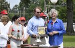 A group of people stand outdoors, one man holding a framed award, others holding red roses, gathered around a table with a bell.
