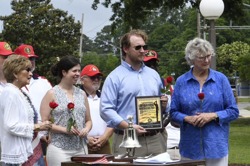 A group of people at an outdoor event hold red roses; a man in sunglasses holds a plaque and stands by a bell and table.