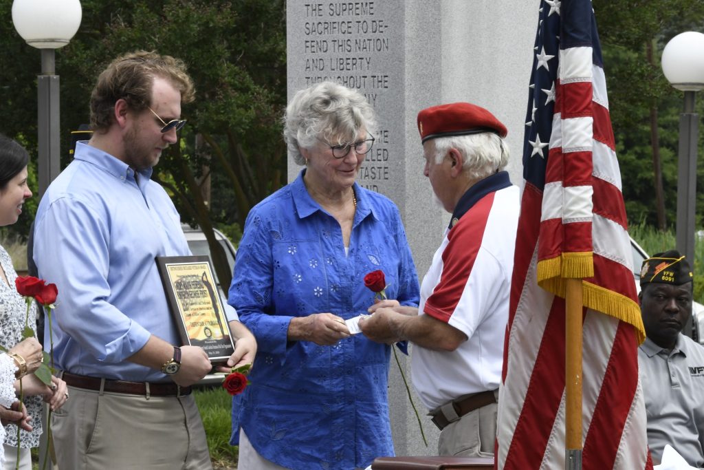 Three people standing by an American flag during a ceremony; one holds a plaque, another receives a rose from a man in a red beret.