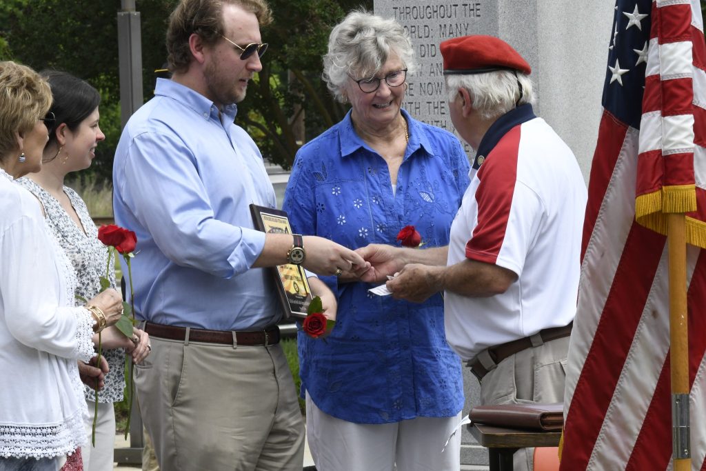 Three people standing outdoors, exchanging items and holding red flowers. One man wears a red hat and white shirt, another holds a plaque, and a woman stands between them.