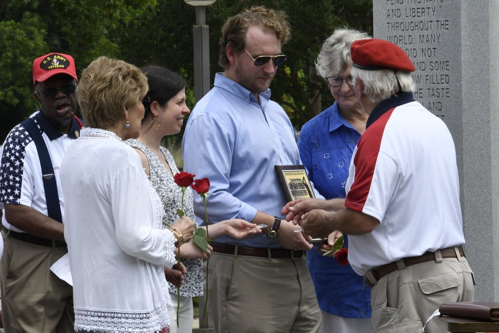 A group of people standing outdoors, some holding red roses, while one person hands a plaque to another in a casual, respectful gathering.