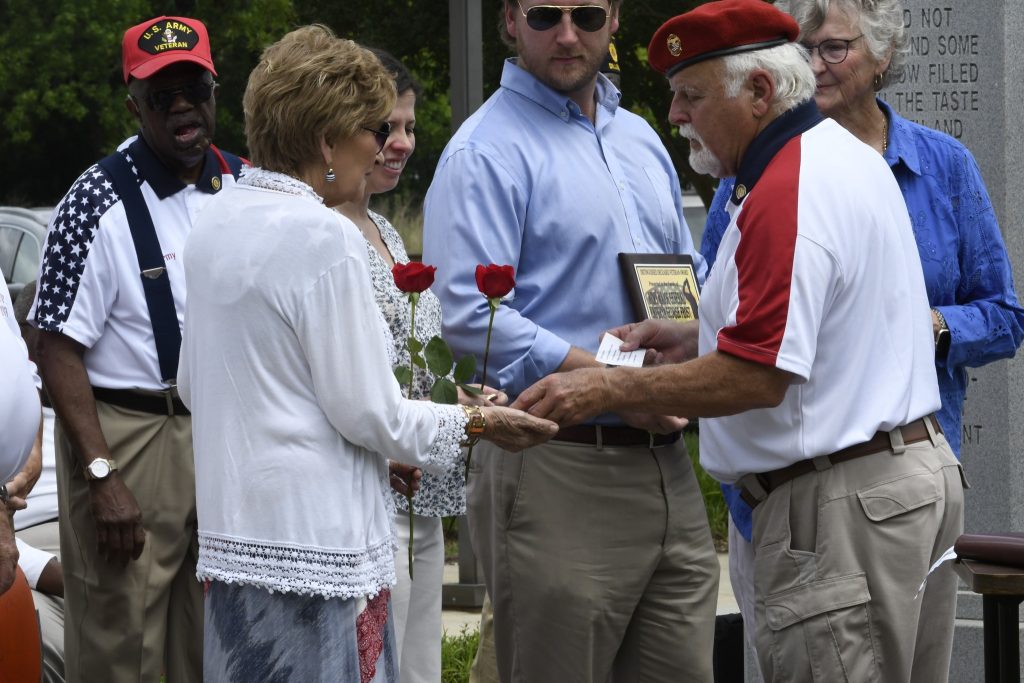A group of people gathered outdoors, one woman holding red roses, while an older man in a red beret hands her an item, others watch.