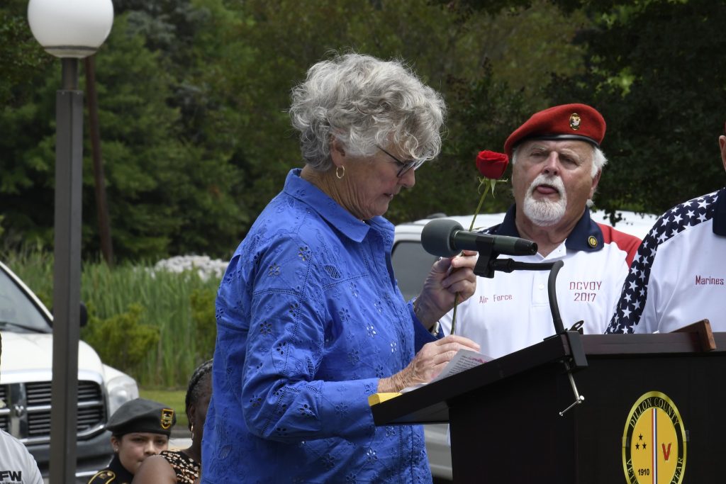 older woman speaks at a podium outdoors while a man in a white uniform and red beret holds a red flower beside her.