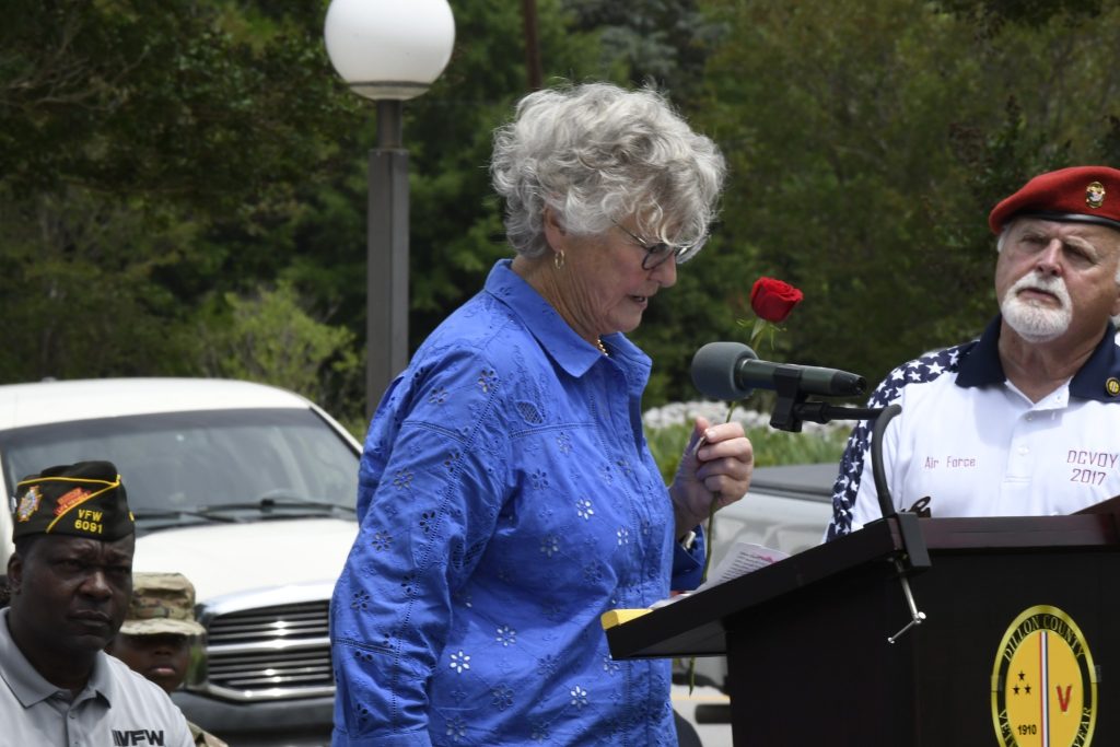 older woman with curly gray hair speaks at an outdoor podium, wearing a blue shirt. A car and greenery are visible in the background.