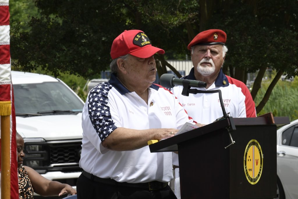 Two men wearing red caps and white shirts with patriotic designs stand outdoors; one speaks at a podium with a microphone, the other stands behind him.