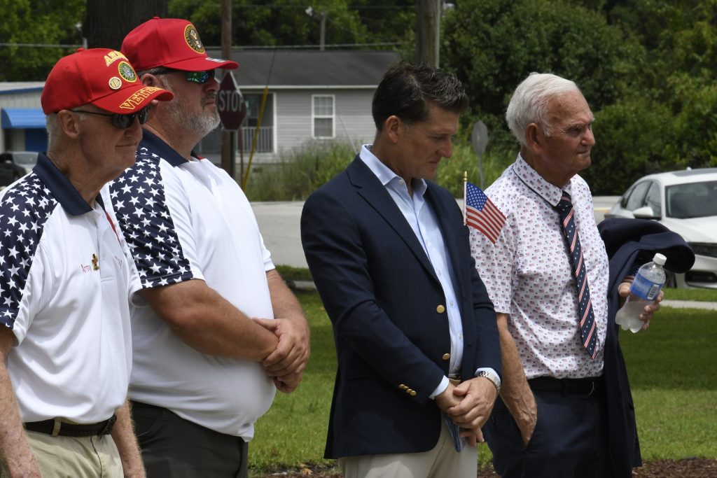 Four men standing outdoors, two wearing patriotic shirts and hats, one in a suit holding a small American flag, participating in what appears to be a memorial or patriotic event.