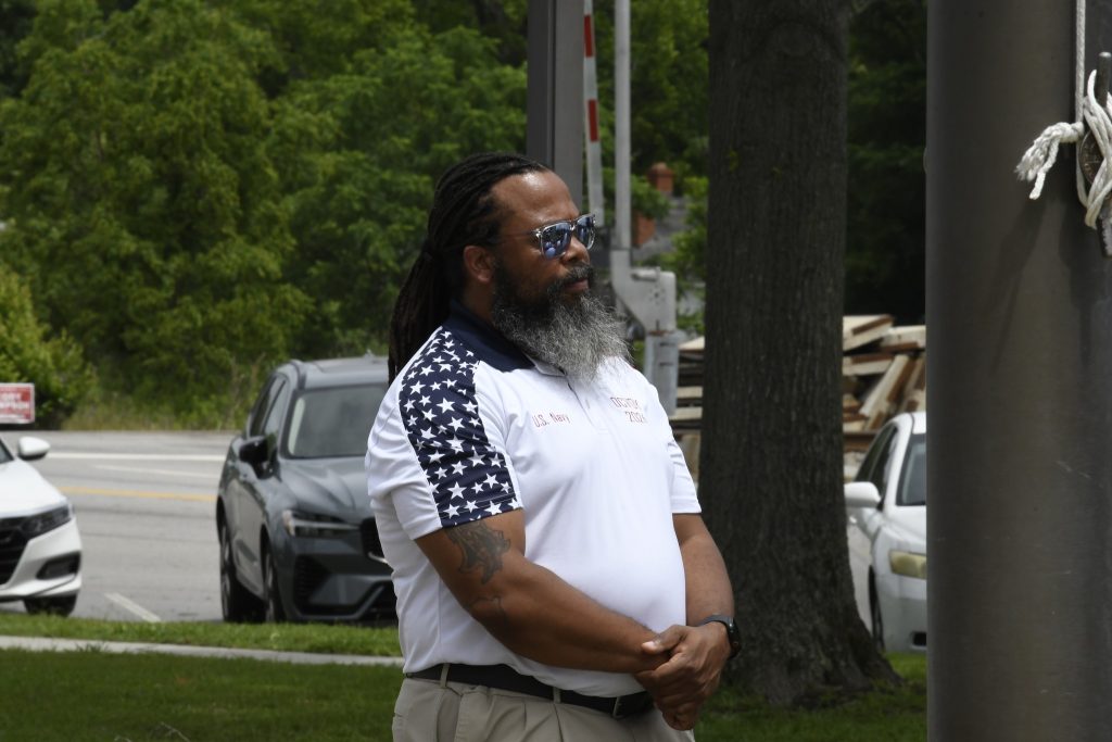 A man with sunglasses and a white shirt with star patterns stands outdoors, with cars and trees visible in the background.
