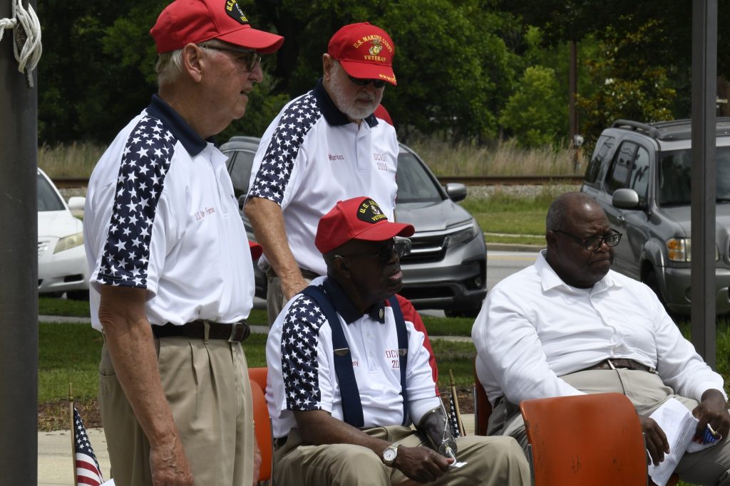Four older men sit and stand outdoors, three wearing matching patriotic shirts and red caps, attending an event on a sunny day.