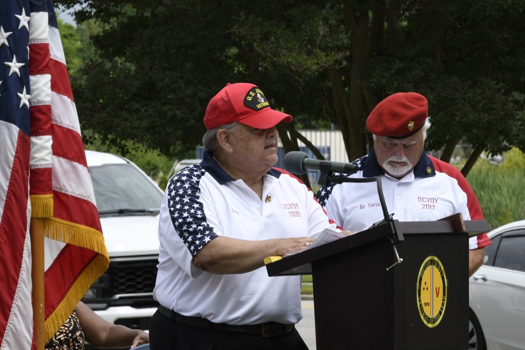 Two men in patriotic shirts and red hats stand at a podium outdoors, with one speaking and the other holding papers. An American flag is partially visible.