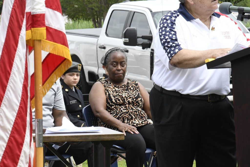 People seated outdoors near a table and an American flag, with a white truck in the background. One person stands in the foreground, partially out of frame.