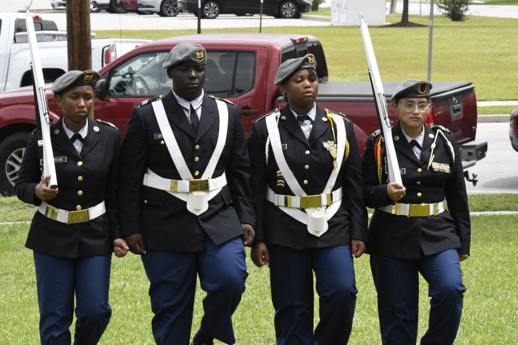 Four uniformed cadets march in formation outdoors, with one holding a ceremonial sword. A red pickup truck and grassy area are visible in the background.