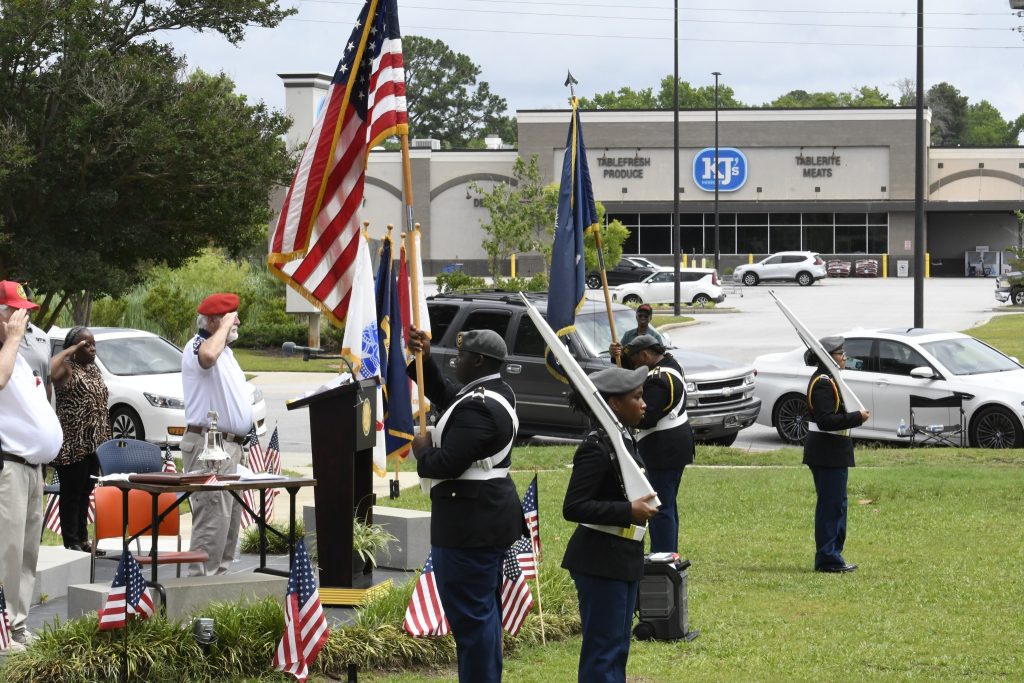 Uniformed honor guard holds swords and flags during an outdoor ceremony, with American flags and a man saluting near a podium, cars and buildings in the background.