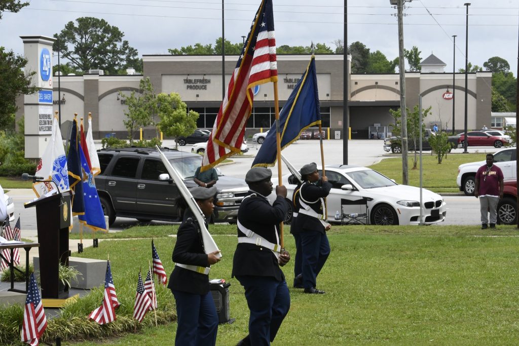 Uniformed honor guard carries American and other flags in a grassy area with small flags, nearby parked cars, and buildings in the background.