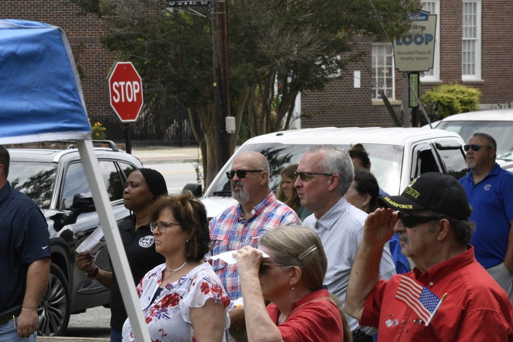 A group of people standing outdoors in front of a white van, some shielding their eyes, with a stop sign and trees in the background.