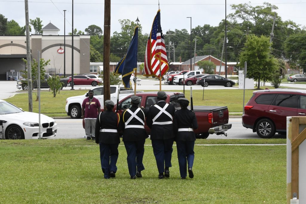 Four uniformed individuals carry American and blue flags, walking on grass, with cars and buildings in the background.