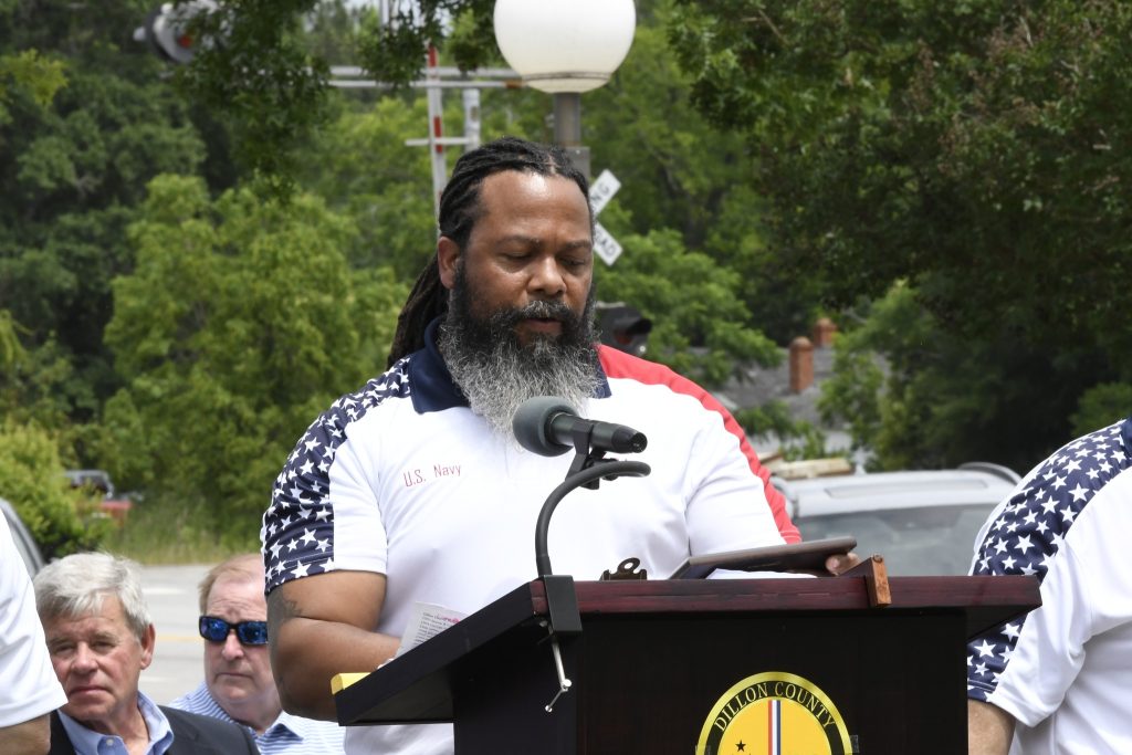 Man with a long beard speaks at an outdoor podium during a public event, with trees and people visible in the background.