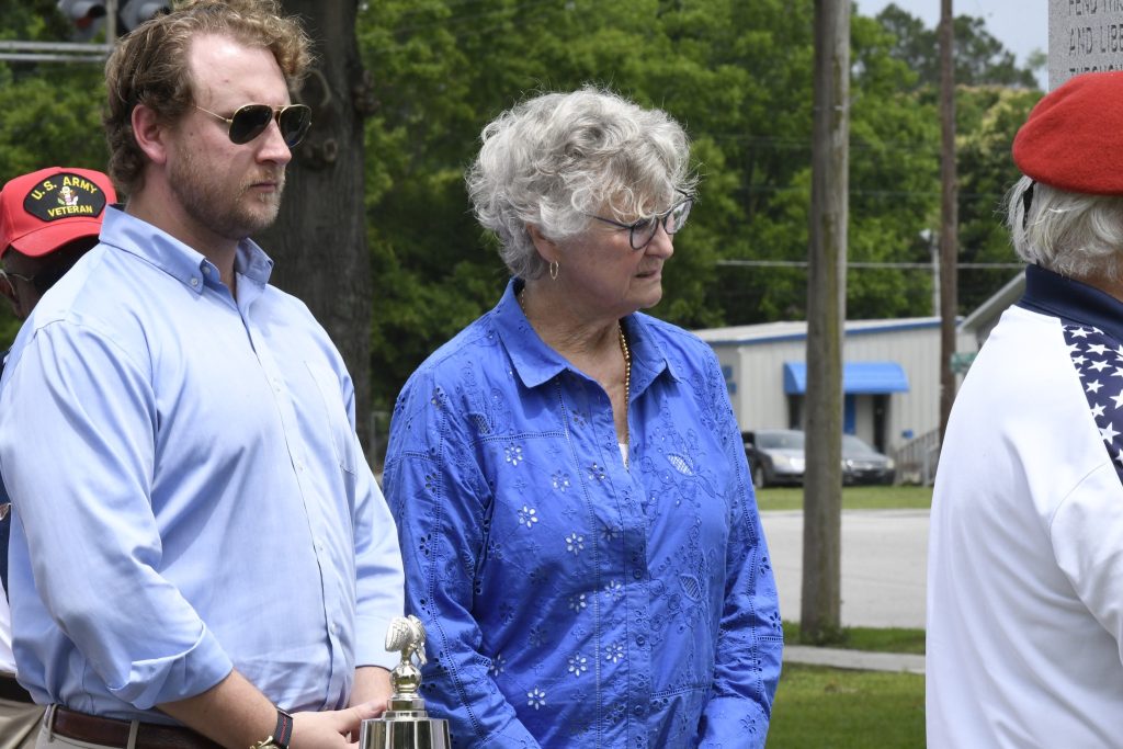 A man and an older woman stand outdoors side by side, both wearing blue shirts, with trees and a building in the background.