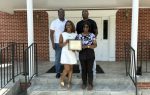 Four people stand on stone steps outside a brick building; one woman in front holds a framed certificate, and they are all smiling for the camera.