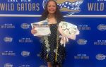 Smiling student in polka dot dress holds bouquet, certificate, and graduation cap, standing in front of Lake View Wild Gators backdrop.