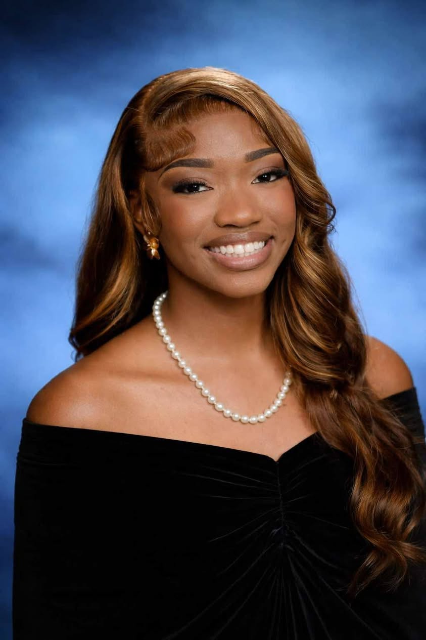 Young woman with long brown hair, wearing an off-shoulder black top and pearl necklace, smiling in front of a blue studio background.