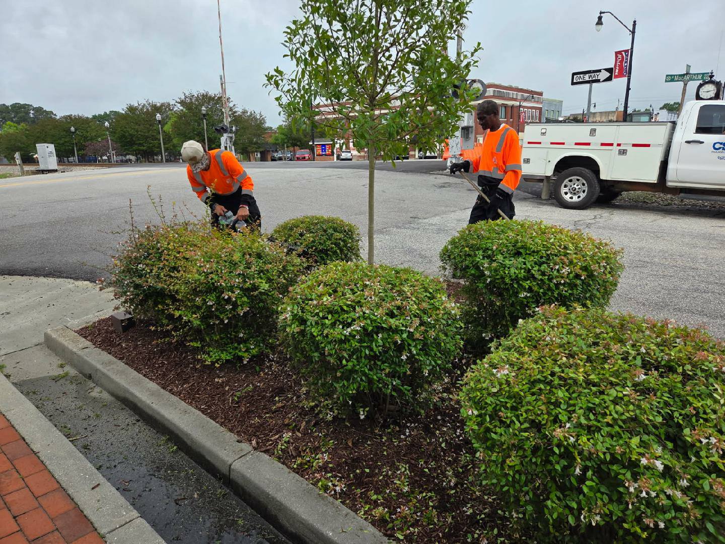 Two workers in orange shirts tending to bushes and landscaping in a parking lot, with a utility truck in the background.