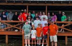A large group of people of various ages stand together on a wooden porch, posing for a group photo outdoors under a cloudy sky.
