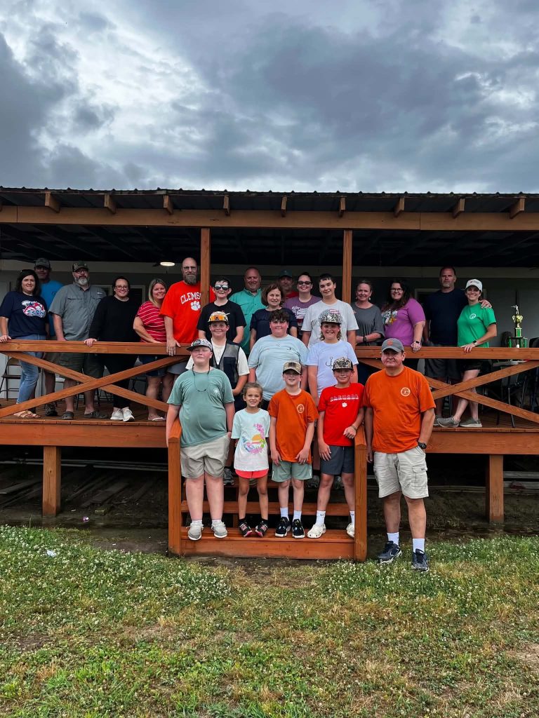 A large group of people of various ages stand together on a wooden porch, posing for a group photo outdoors under a cloudy sky.