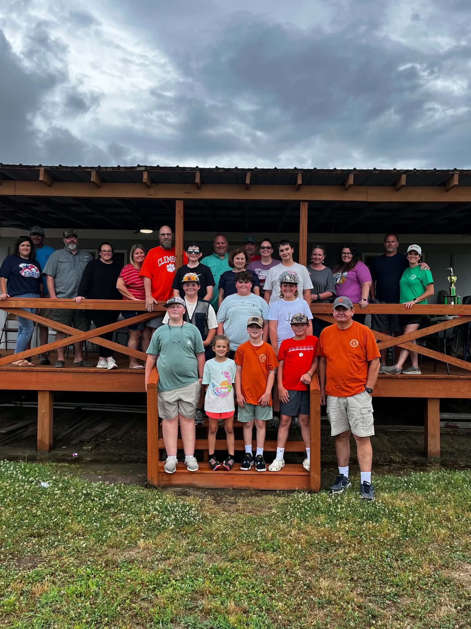 A large group of people of various ages stand together on a wooden porch, posing for a group photo outdoors under a cloudy sky.
