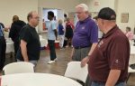 Three men stand and talk near a table inside a community center, with several other people visible in the background.