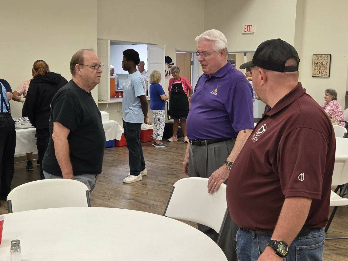 Three men stand and talk near a table inside a community center, with several other people visible in the background.