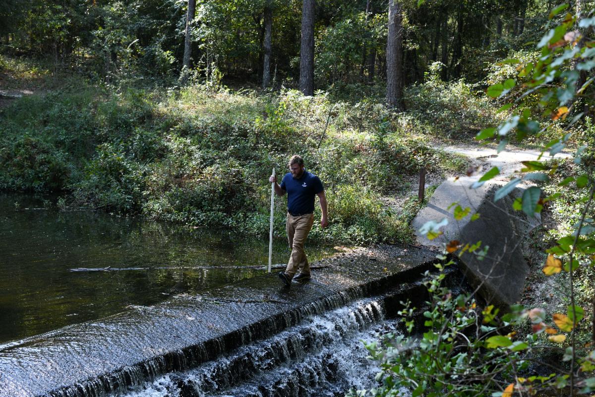 A person stands on a small concrete dam holding a stick, surrounded by flowing water and dense trees in a forested area.