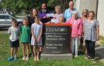 A group of people, including children and adults, stand smiling around a Clemson sign for Dillon County Extension Office, outdoors on a sunny day.
