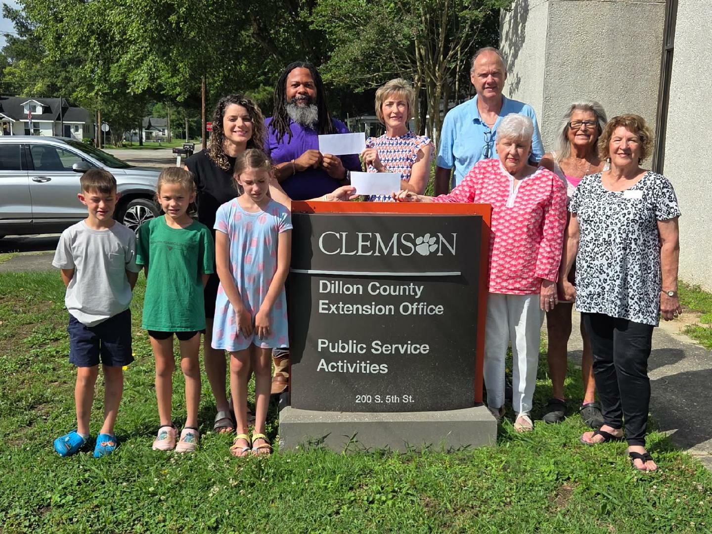 A group of people, including children and adults, stand smiling around a Clemson sign for Dillon County Extension Office, outdoors on a sunny day.