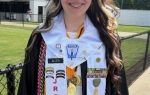 Young woman in graduation gown and stole stands outdoors, smiling, with ROTC and honor cords, medals, and pins displayed. Green trees and buildings in background.