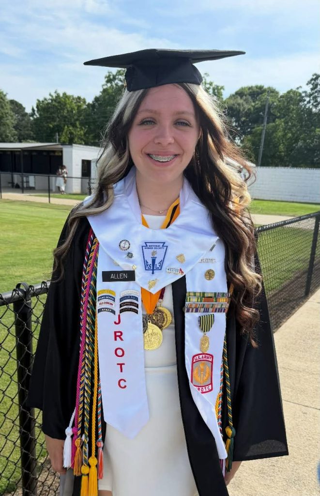 Young woman in graduation gown and stole stands outdoors, smiling, with ROTC and honor cords, medals, and pins displayed. Green trees and buildings in background.