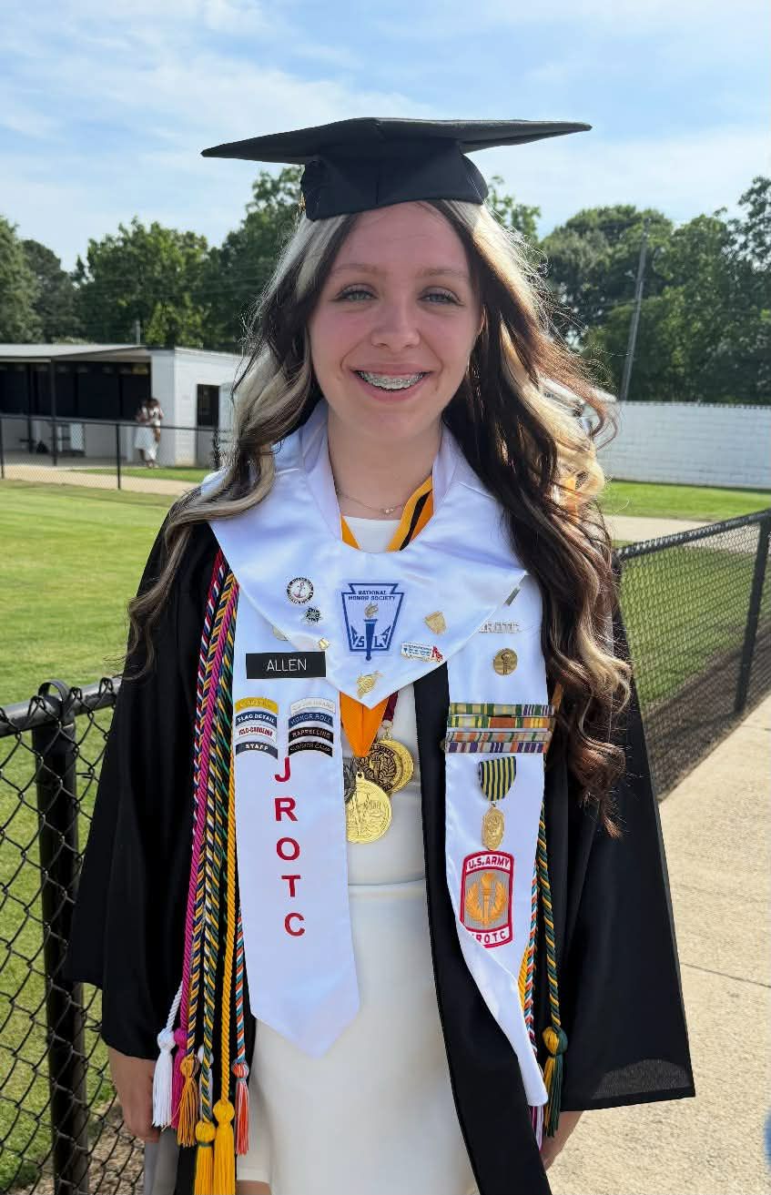 Young woman in graduation gown and stole stands outdoors, smiling, with ROTC and honor cords, medals, and pins displayed. Green trees and buildings in background.