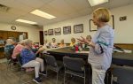A group of elderly women sit around a table in a meeting room while one woman stands and speaks to them.