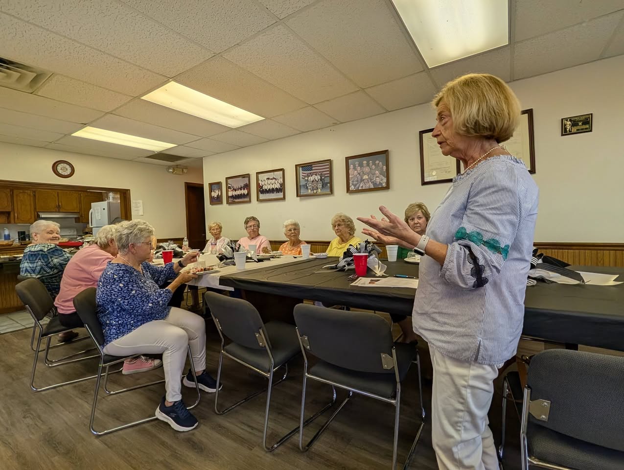 A group of elderly women sit around a table in a meeting room while one woman stands and speaks to them.