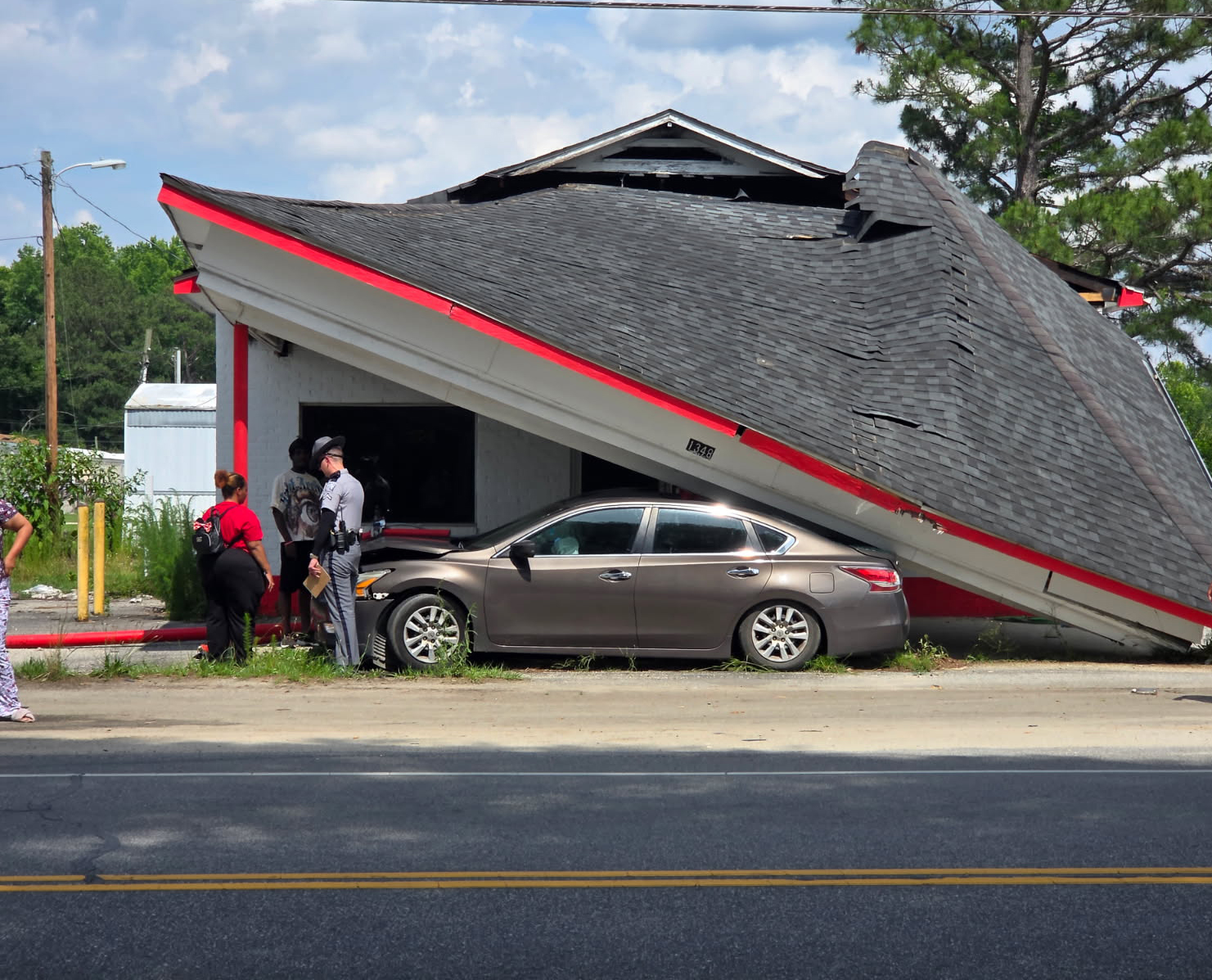 Collapsed building roof on top of a parked car, with several people standing nearby on the sidewalk, observing the damage.