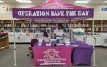 Two people stand behind a purple booth and canopy reading "OPERATION SAVE THE DAY" inside a library, promoting an organization with pamphlets and informational materials.