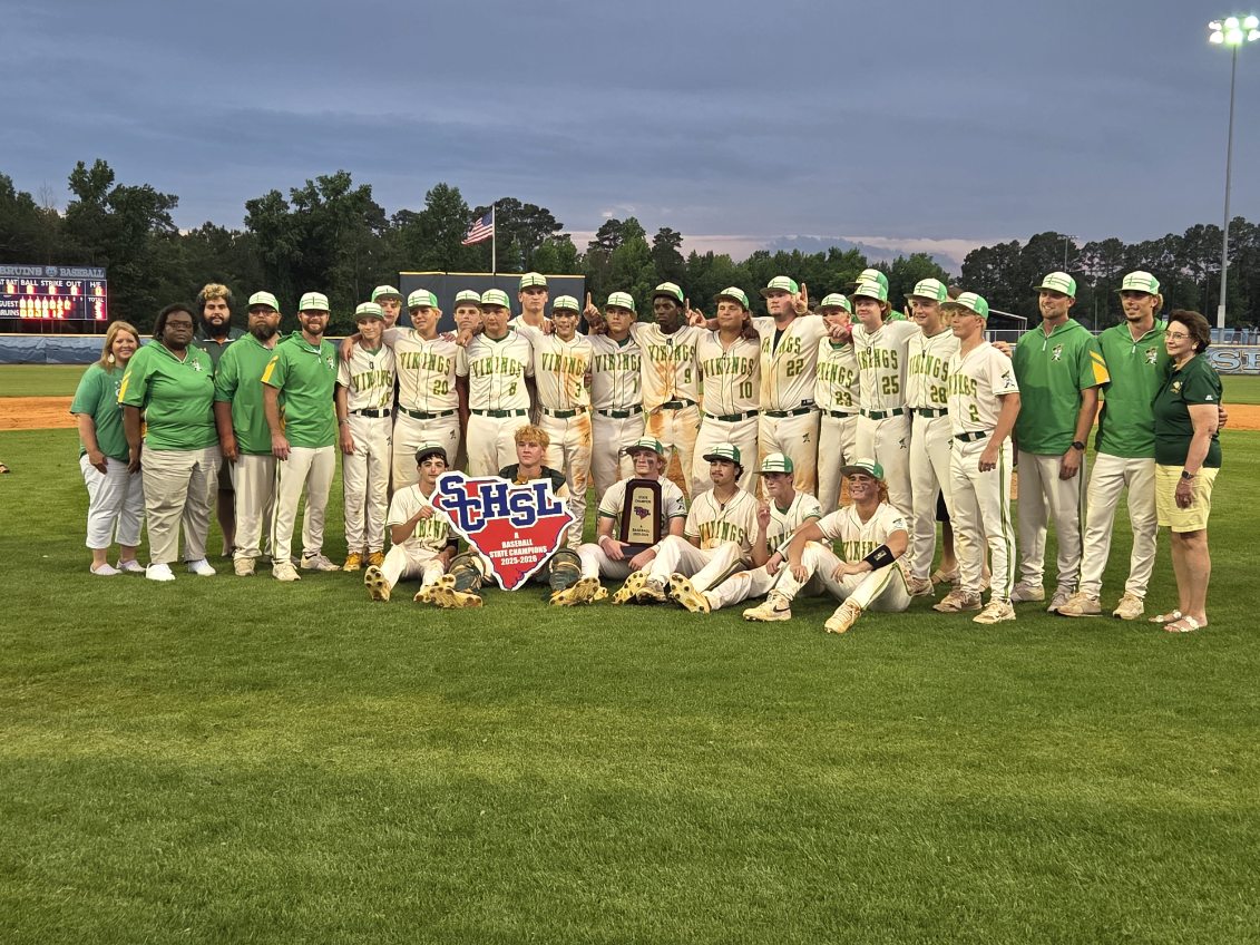 A high school baseball team poses together on a field, holding a state championship sign, with coaches in green shirts standing beside them.