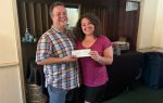 Two people smiling and standing indoors, holding a check together.