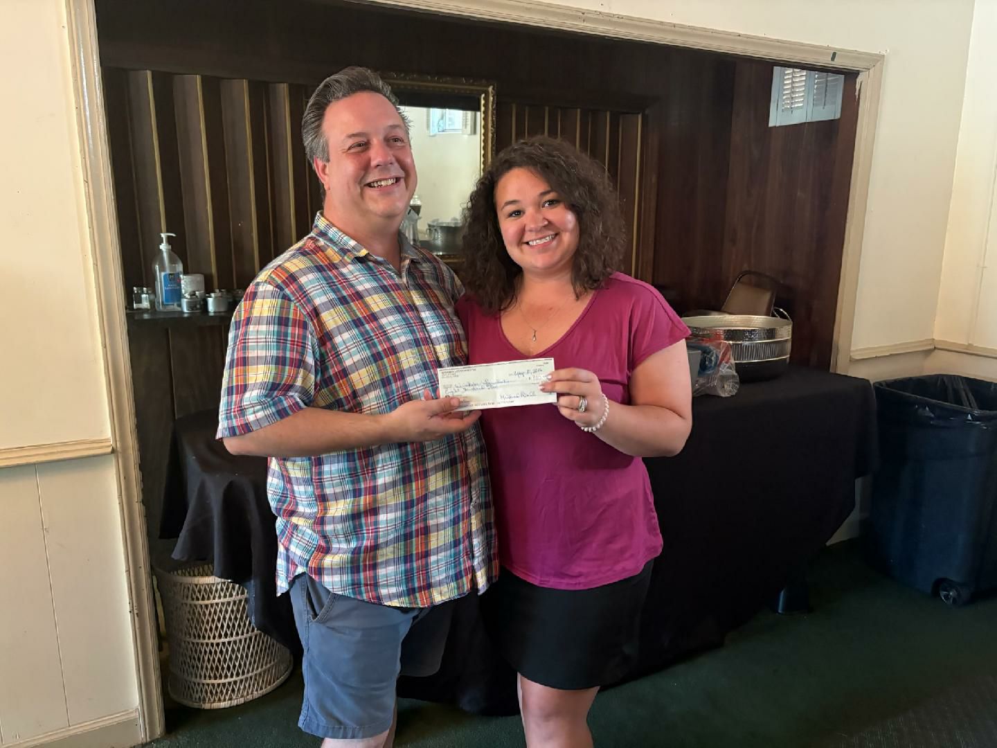 Two people smiling and standing indoors, holding a check together.