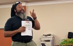 A man stands indoors holding a printed sheet, gesturing with his other hand, wearing a black shirt and cap, with a copier and table visible behind him.